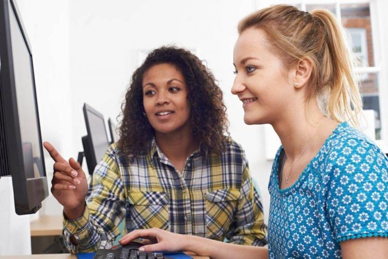 duas mulheres sentadas em uma mesa olhando para a tela de um computador