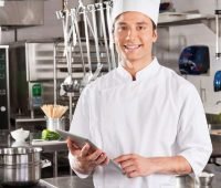um homem com chapéu de chef segurando um tablet em uma cozinha