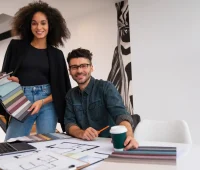 Profissional de design de interiores e um colega em reunião de projeto, analisando mostruários de cores e plantas arquitetônicas em um ambiente de escritório moderno.