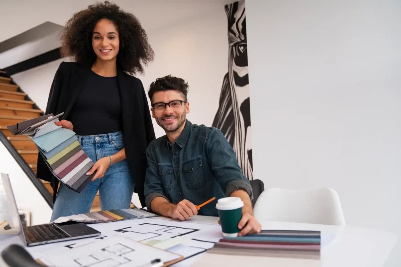 Profissional de design de interiores e um colega em reunião de projeto, analisando mostruários de cores e plantas arquitetônicas em um ambiente de escritório moderno.