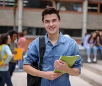 Jovem estudante sorridente com mochila, segurando caderno em ambiente escolar, rodeado de colegas, representando a vida acadêmica.