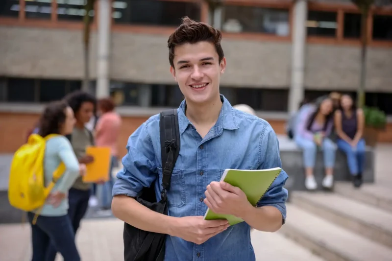 Jovem estudante sorridente com mochila, segurando caderno em ambiente escolar, rodeado de colegas, representando a vida acadêmica.
