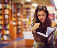 Jovem mulher lendo um livro em uma biblioteca, concentrada e rodeada por estantes cheias de livros, ideal para amantes da leitura.