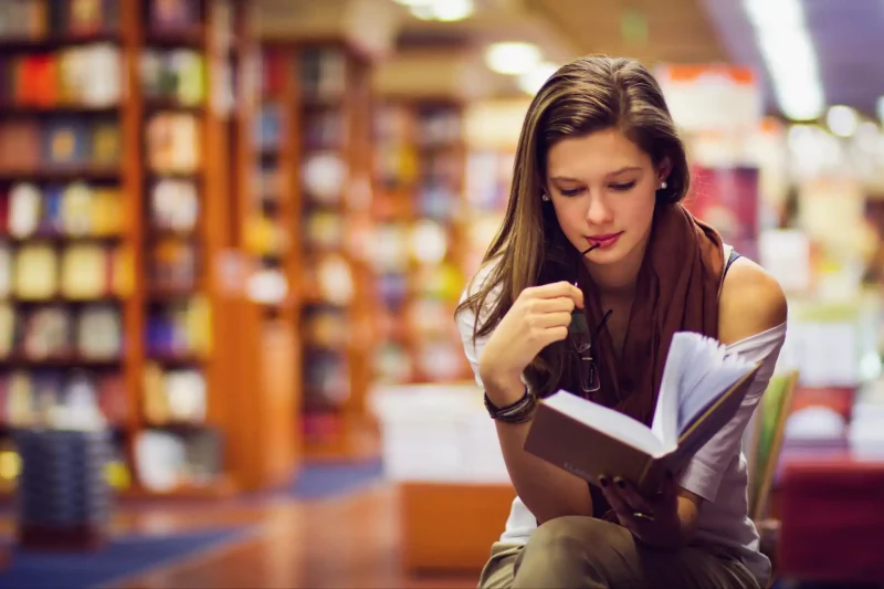 Jovem mulher lendo um livro em uma biblioteca, concentrada e rodeada por estantes cheias de livros, ideal para amantes da leitura.