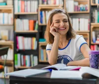 Estudante do Enem sorridente estudando em biblioteca com livros ao redor, preparando para a prova do exame nacional com foco e otimismo.