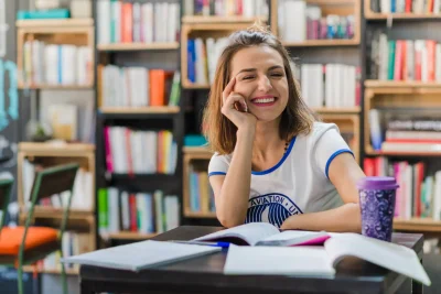 Estudante do Enem sorridente estudando em biblioteca com livros ao redor, preparando para a prova do exame nacional com foco e otimismo.