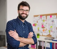 Homem sorridente com barba e óculos, representando gestão de projetos, em ambiente de escritório com quadro de anotações e documentos.