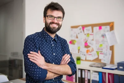 Homem sorridente com barba e óculos, representando gestão de projetos, em ambiente de escritório com quadro de anotações e documentos.
