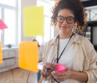 Mulher jovem de óculos, com cabelo cacheado, escrevendo em um planner rosa em ambiente de escritório com post-its coloridos na parede, promovendo organização, produtividade e planejamento.