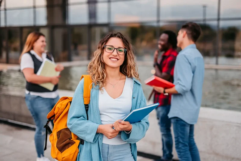 Estudantes felizes interagindo ao ar livre, com materiais escolares como cadernos e mochilas coloridas. Um ambiente universitário vibrante e colaborativo.