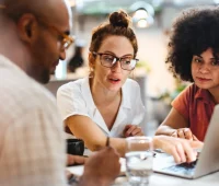 Grupo de pessoas discutindo ideias em um ambiente de trabalho colaborativo, focadas em um laptop e com anotações visíveis.