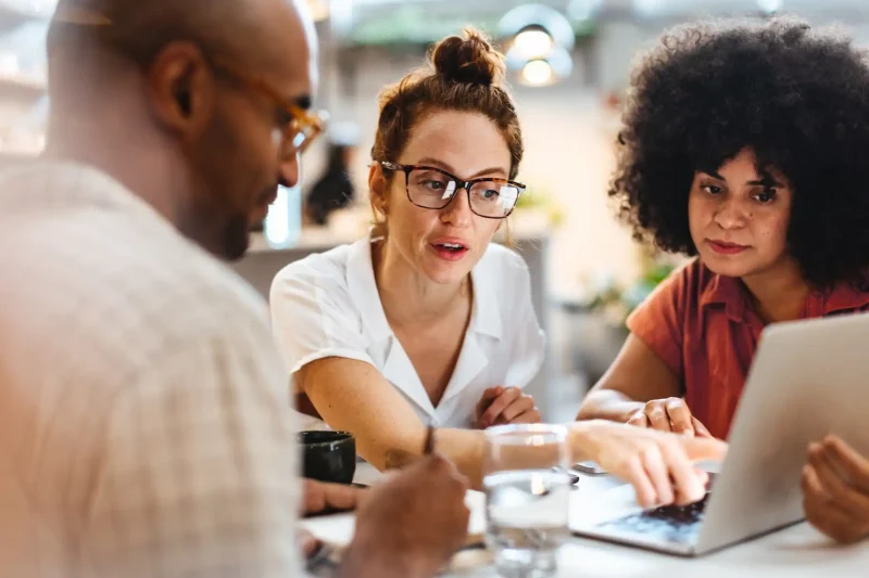 Grupo de pessoas discutindo ideias em um ambiente de trabalho colaborativo, focadas em um laptop e com anotações visíveis.