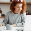 Mulher com cabelo cacheado estudando ou trabalhando em casa. Ela usa um suéter cinza, está concentrada diante de um laptop, escrevendo anotações.