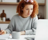 Mulher com cabelo cacheado estudando ou trabalhando em casa. Ela usa um suéter cinza, está concentrada diante de um laptop, escrevendo anotações.