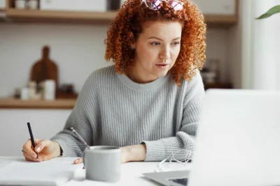 Mulher com cabelo cacheado estudando ou trabalhando em casa. Ela usa um suéter cinza, está concentrada diante de um laptop, escrevendo anotações.