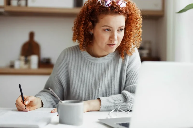 Mulher com cabelo cacheado estudando ou trabalhando em casa. Ela usa um suéter cinza, está concentrada diante de um laptop, escrevendo anotações.