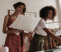 Duas mulheres sorridentes revisando desenhos e projetos em um ambiente de trabalho criativo, com fundo de parede branca e decoração moderna.