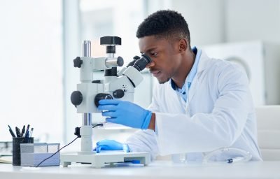 Cientista realizando pesquisa com um microscópio em laboratório, usando jaleco branco e luvas azuis.