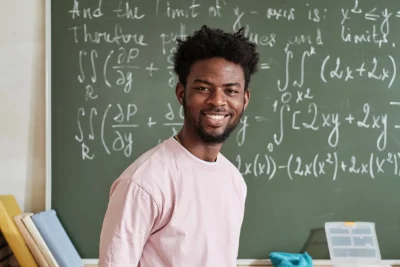Professor de matemática sorridente em sala de aula, com quadro cheio de equações no fundo, representando ensino de matemática e matemática básica.