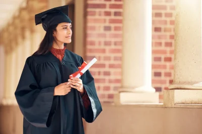 Mulher em trajes de formatura segurando diploma, olhando para o horizonte. A imagem representa a celebração da conquista acadêmica e realizações.
