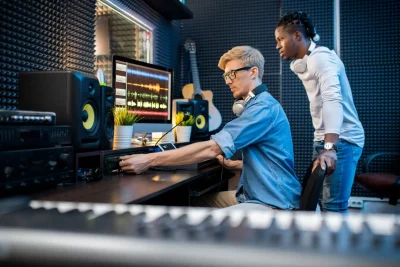 Dois profissionais trabalhando em um estúdio de gravação, focando na produção musical, com equipamentos modernos ao fundo.