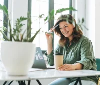 Mulher sorridente trabalhando em casa, com laptop sobre a mesa, segurando uma caneta e um copo de café, rodeada por plantas verdes.