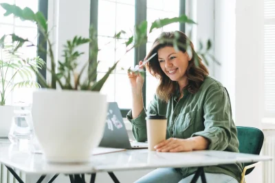 Mulher sorridente trabalhando em casa, com laptop sobre a mesa, segurando uma caneta e um copo de café, rodeada por plantas verdes.
