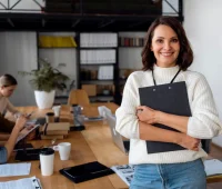 Mulher sorridente em ambiente de escritório moderno, segurando pastas e com colegas de trabalho ao fundo, em uma reunião de equipe.