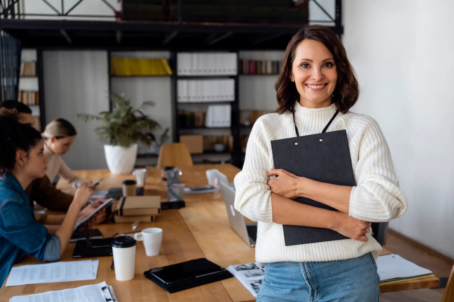 Mulher sorridente em ambiente de escritório moderno, segurando pastas e com colegas de trabalho ao fundo, em uma reunião de equipe.