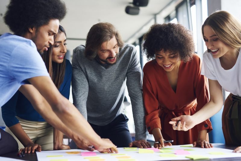 Grupo diverso de pessoas colaborando em atividades de brainstorming com notas adesivas coloridas em uma mesa, promovendo trabalho em equipe e criatividade.