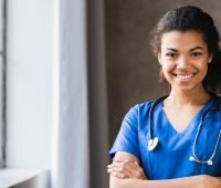 Mulher médica sorrindo, usando uniforme azul e estetoscópio ao redor do pescoço, em ambiente clínico claro e organizado.
