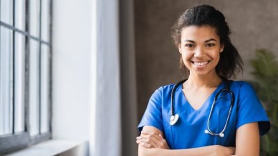 Mulher médica sorrindo, usando uniforme azul e estetoscópio ao redor do pescoço, em ambiente clínico claro e organizado.