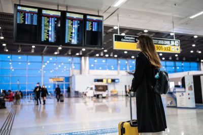 Mulher assistindo ao painel de voos em um aeroporto, com mala amarela ao seu lado, visivelmente aguardando informações sobre seu voo.