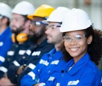 Imagem de um grupo diversificado de trabalhadores usando capacetes de segurança e óculos de proteção, representando a segurança no trabalho e a força da mão de obra.