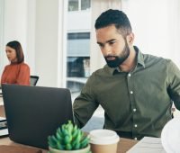 Homem usando laptop em ambiente de escritório moderno, concentrado no trabalho enquanto mulher ao fundo também trabalha.