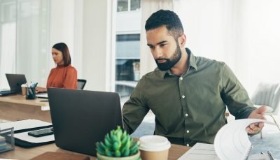 Homem usando laptop em ambiente de escritório moderno, concentrado no trabalho enquanto mulher ao fundo também trabalha.