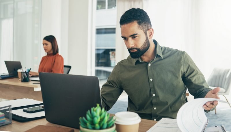 Homem usando laptop em ambiente de escritório moderno, concentrado no trabalho enquanto mulher ao fundo também trabalha.