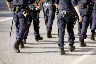 Imagem de policiais em uniforme, caminhando em direção ao alvo. A cena demonstra a atuação das forças de segurança pública em um ambiente urbano.