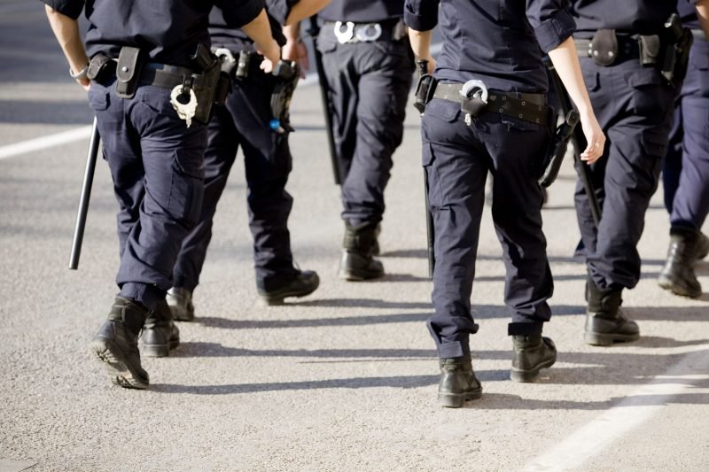 Imagem de policiais em uniforme, caminhando em direção ao alvo. A cena demonstra a atuação das forças de segurança pública em um ambiente urbano.