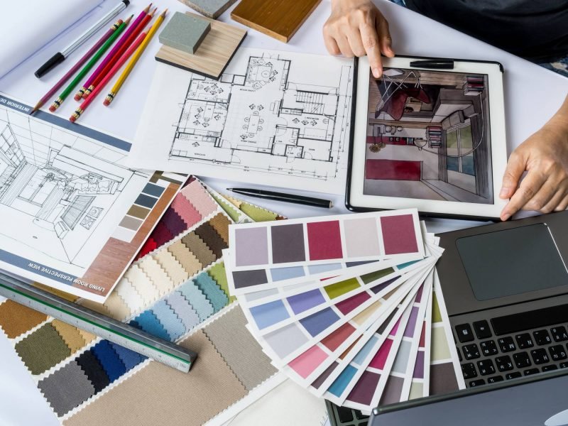 Imagem de um projeto de design de interiores com amostras de cores, papel de croqui, lápis de cor e um tablet exibindo um ambiente decorado.