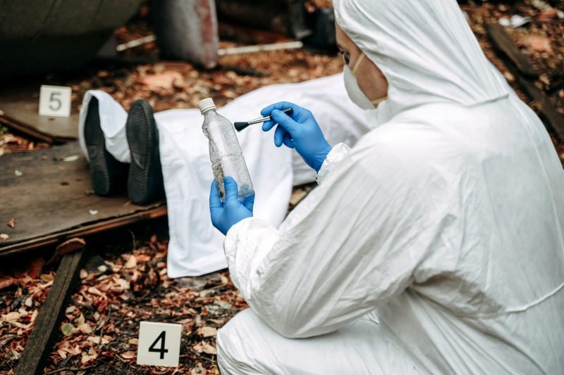 Profissional forense analisando evidências em cena de crime, com botellas e objetos identificados, em um cenário misterioso.