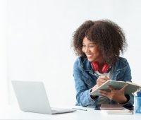 Mulher estudante sorrindo, usando fones de ouvido, estudando com um caderno e notebook em um ambiente moderno. Ideal para motivação nos estudos.