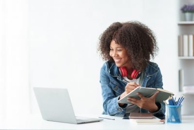 Mulher estudante sorrindo, usando fones de ouvido, estudando com um caderno e notebook em um ambiente moderno. Ideal para motivação nos estudos.