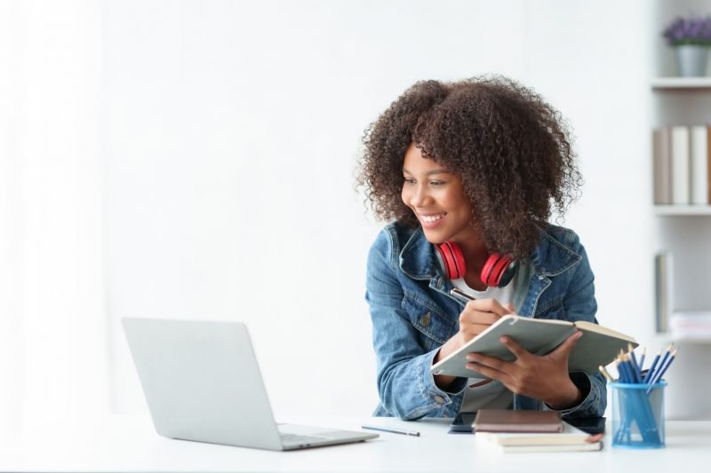 Mulher estudante sorrindo, usando fones de ouvido, estudando com um caderno e notebook em um ambiente moderno. Ideal para motivação nos estudos.