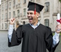 Homem usando beca e capelo comemorando a conquista do certificado Encceja, segurando o diploma com fita vermelha em frente a um prédio universitário.