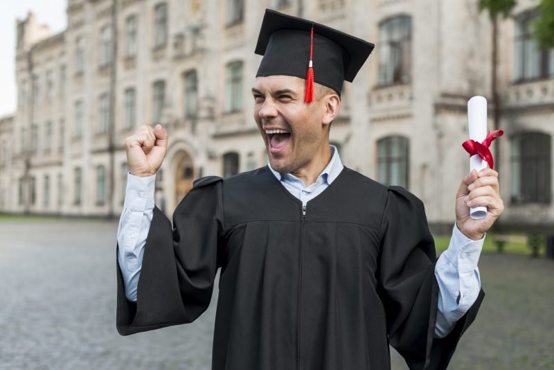 Homem usando beca e capelo comemorando a conquista do certificado Encceja, segurando o diploma com fita vermelha em frente a um prédio universitário.