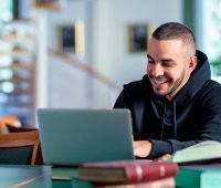 Homem sorridente usando laptop em biblioteca rodeado de livros, concentrado e estudando