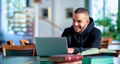 Homem sorridente usando laptop em biblioteca rodeado de livros, concentrado e estudando