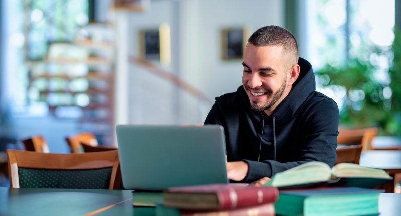 Homem sorridente usando laptop em biblioteca rodeado de livros, concentrado e estudando