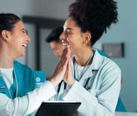 Duas profissionais de saúde sorrindo e se cumprimentando com as mãos em um ambiente de trabalho. A imagem transmite alegria e colaboração no setor de saúde.
